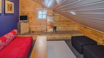 Ein gemütliches Dachzimmer mit einer hölzernen Schräge, einem roten Sofa, einem schwarzen Sessel, einem Holztisch und einem Fernseher auf einer Kommode.