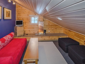 Ett mysigt rum på vinden med ett sluttande tak av trä, en röd soffa, en svart fåtölj, ett träbord och en tv som står på en byrå.