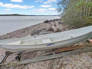 En hvid båd med teksten 'SUMT 420' på skroget, ligger fortøjet ved en grusstrand med stille vand og en fjerntliggende kystlinje i baggrunden.