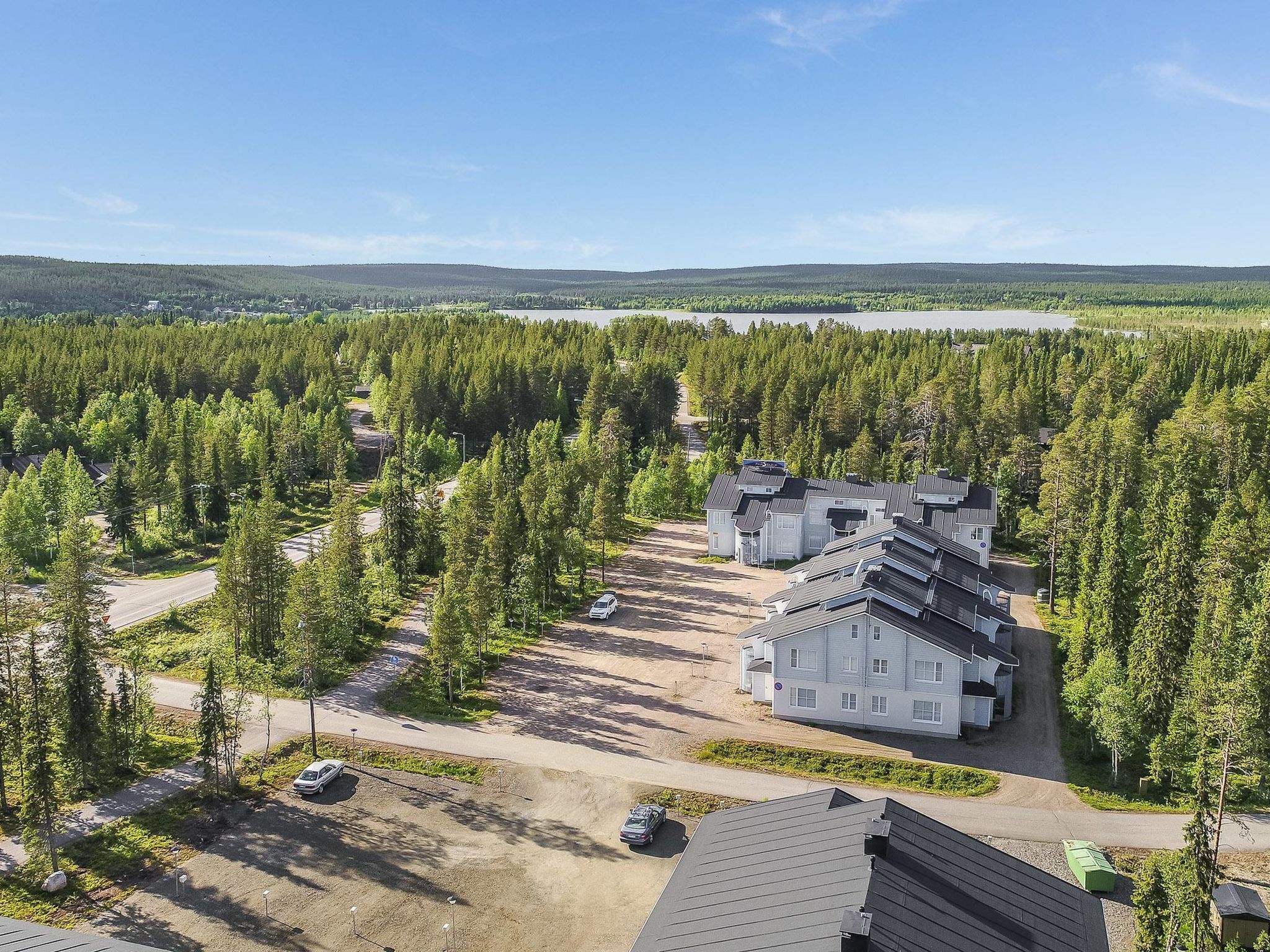 A residential area with multiple houses surrounded by dense forest, a clear sky, and a lake in the background.