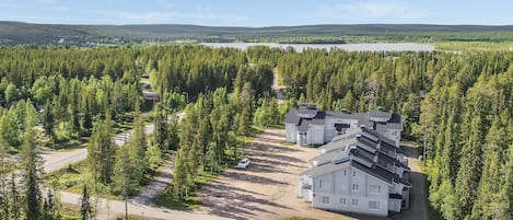 A residential area with multiple houses surrounded by dense forest, a clear sky, and a lake in the background.