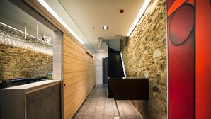 A narrow hallway with a stone wall on one side and a wooden panel wall on the other. A bar area with a counter and glasses is visible on the left. A staircase is seen in the background leading to an upper level. - Hotel Lois (A Coruña)