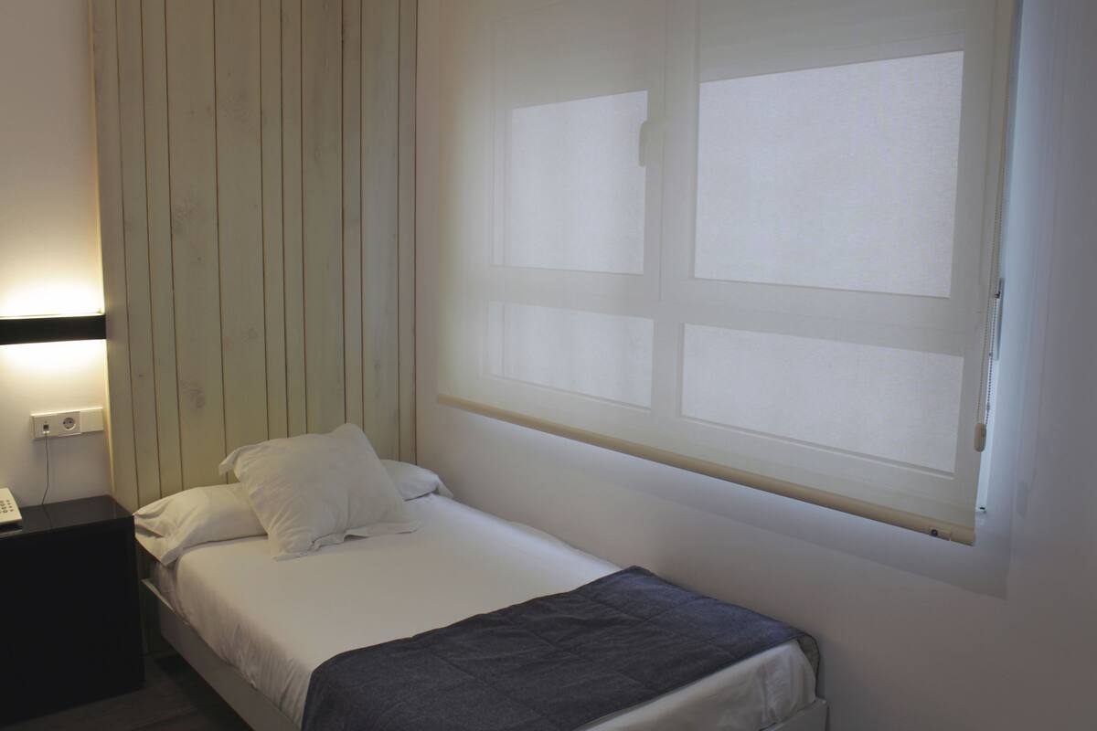 A single bed with a white mattress and gray blanket, a nightstand with a phone, and a window with blinds.