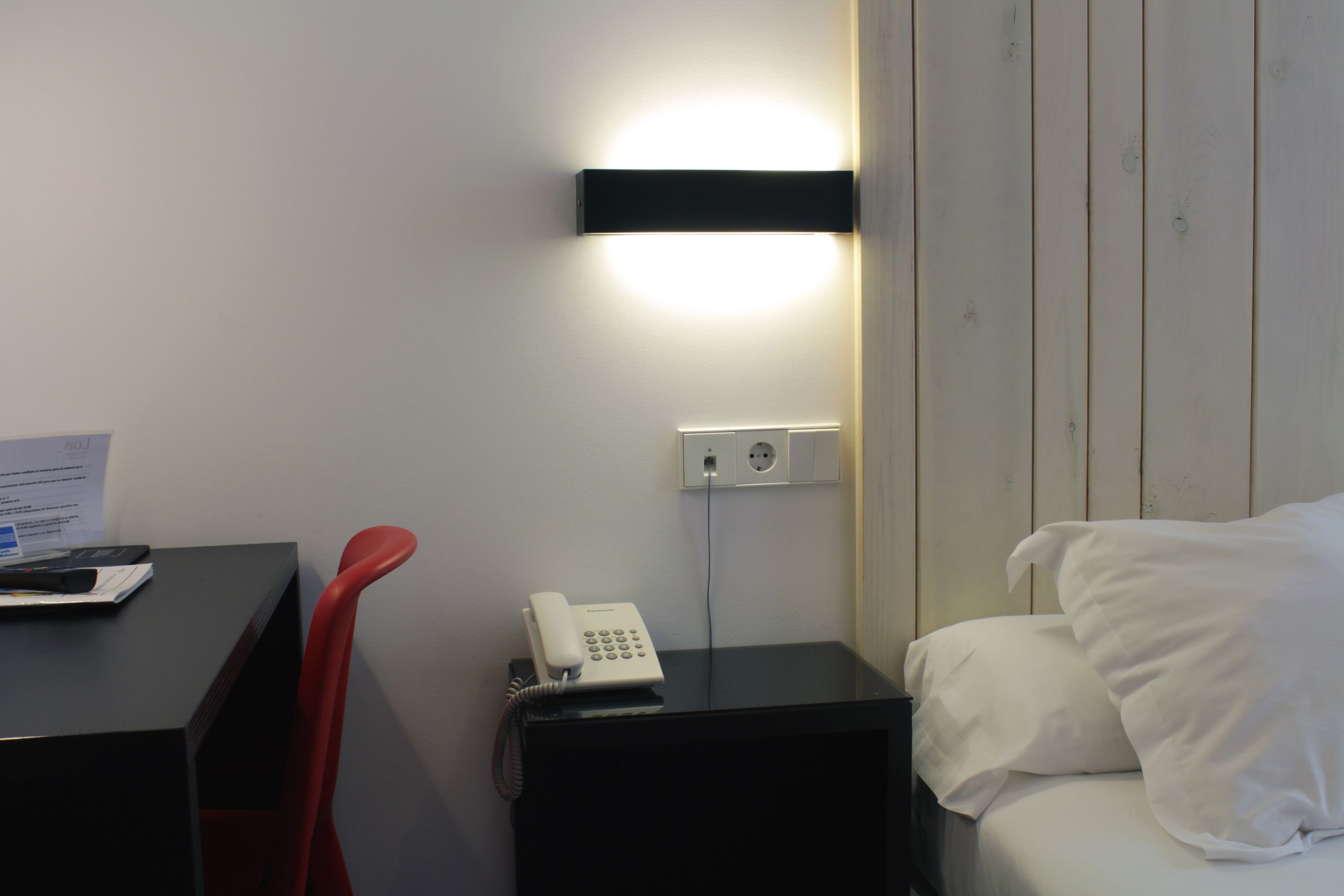Una habitación de hotel con cama, un escritorio con teléfono, y una lámpara de pared.