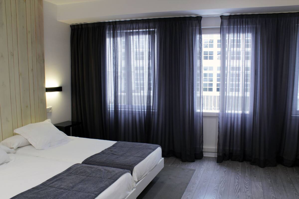 A hotel room with two beds, a large window with dark curtains, a wooden floor, and a nightstand with a lamp.