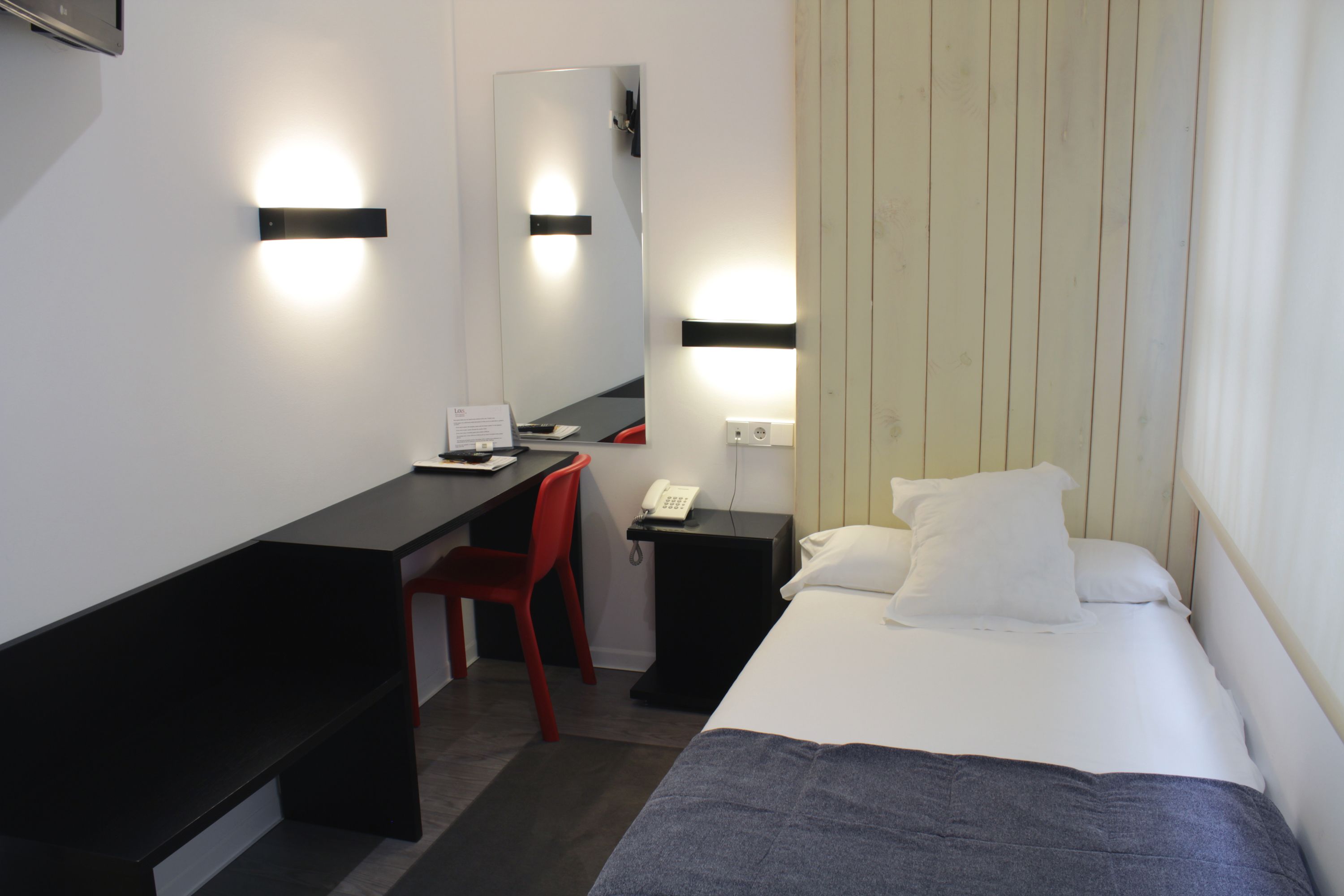 Une chambre d’hôtel comprenant un lit, un bureau, une chaise, une table de chevet avec un téléphone et des lampes fixées au mur.