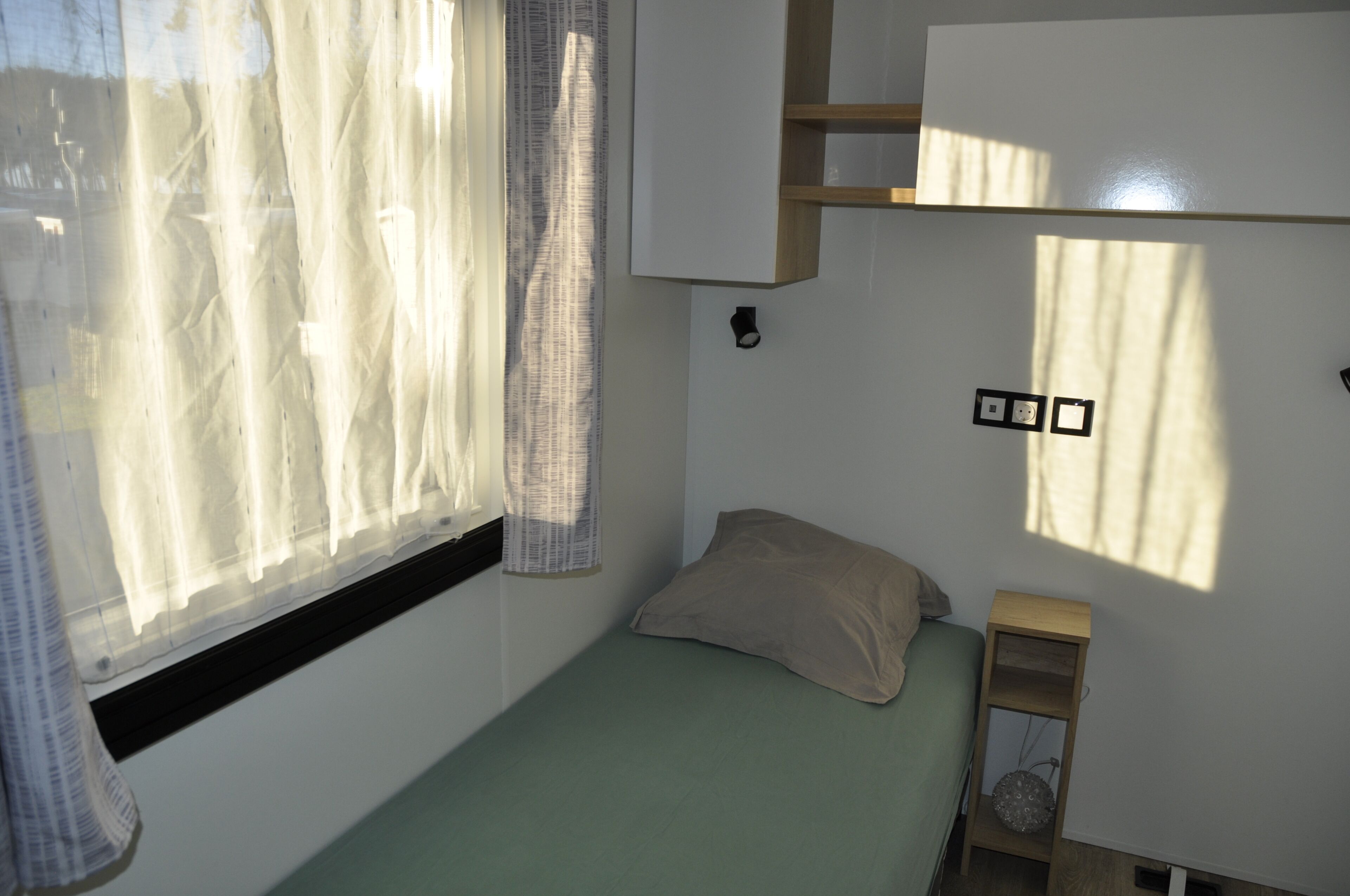 A small, single-bed room with a window, a bedside table, and a wall-mounted shelf.