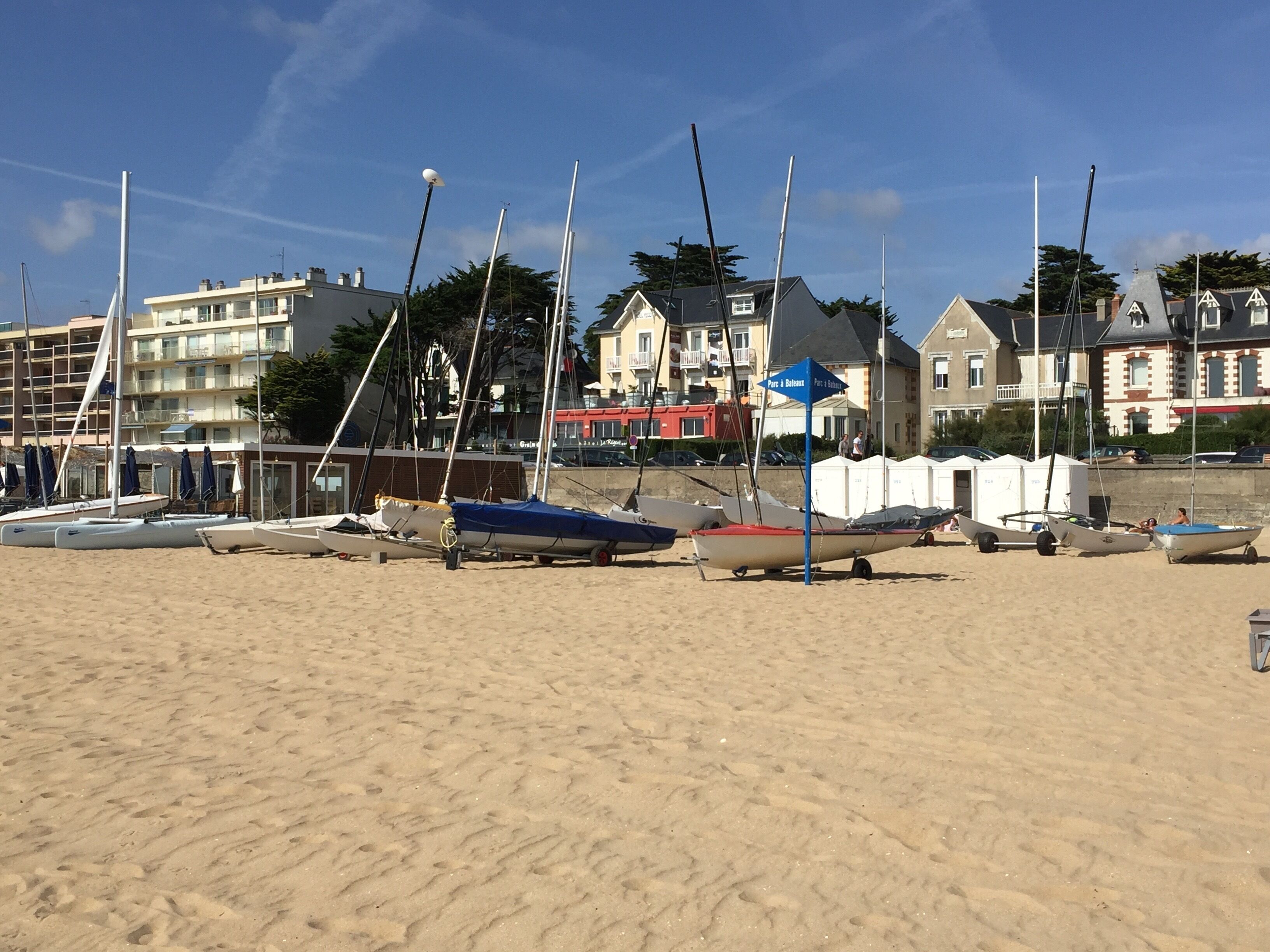 Ein sandiger Strand mit mehreren angestaubten Booten, einem blauen Schild und Gebäuden im Hintergrund.