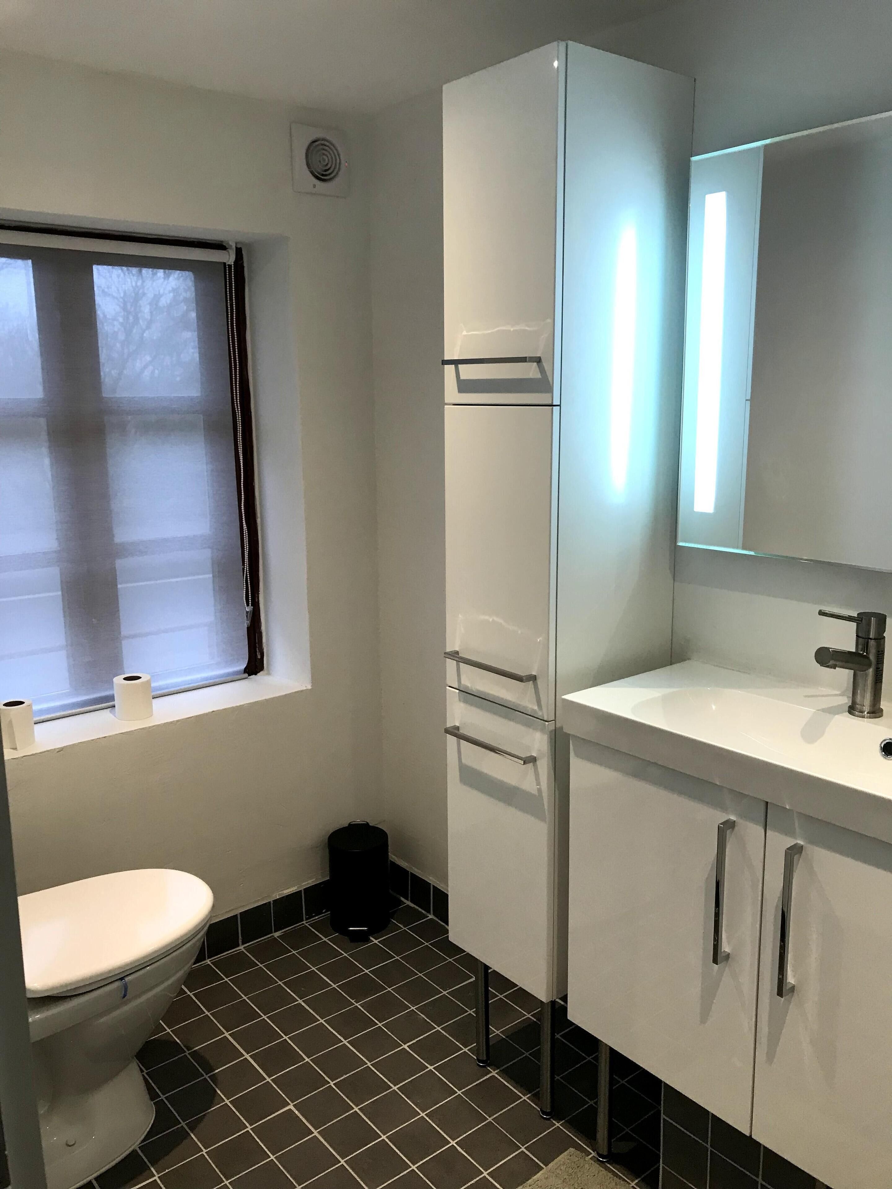 Et badeværelse med toilet, vask med spejl og et vindue med persienner.