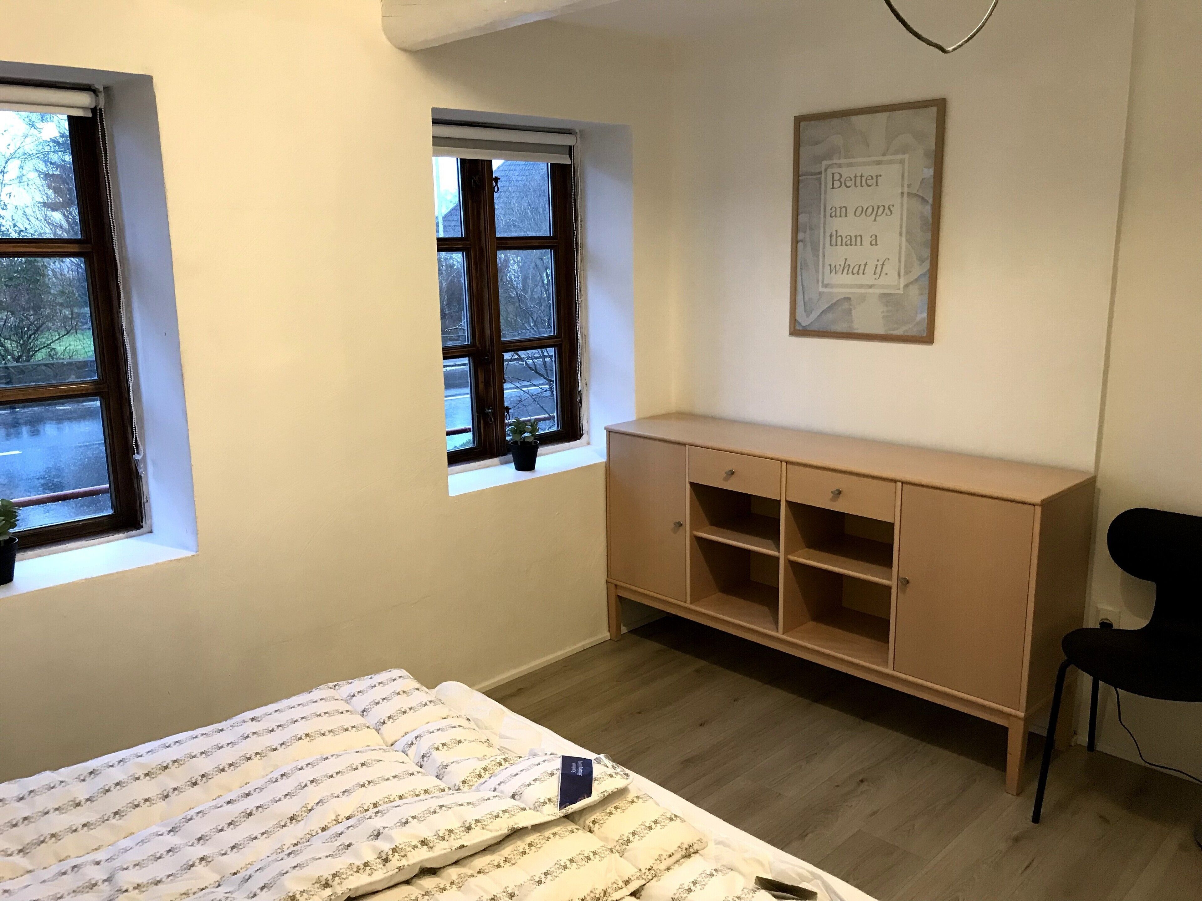A bedroom with two windows, a bed with a white and blue striped duvet, a wooden cabinet, and a black chair.