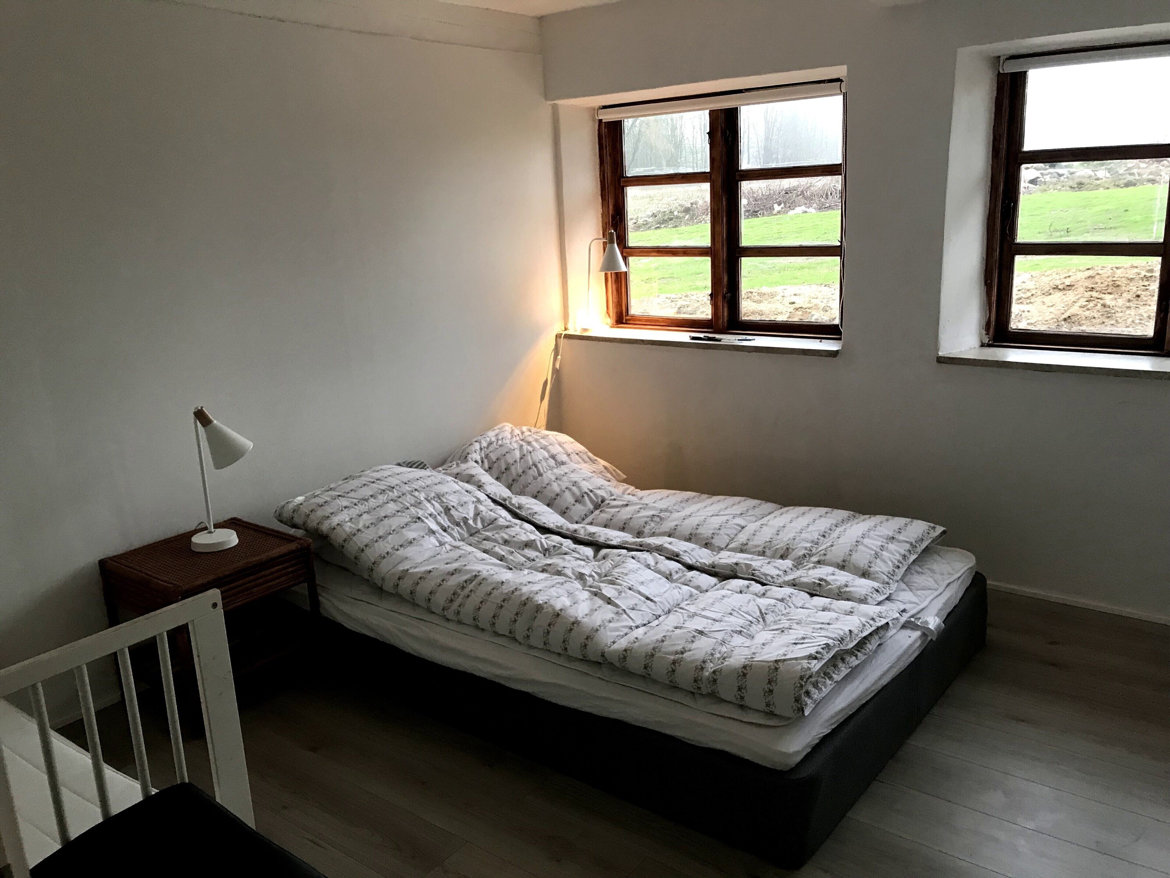 Een kleine kamer met een enkel bed, een houten vloer, een nachtkastje met lamp en twee ramen met uitzicht op een gazon.