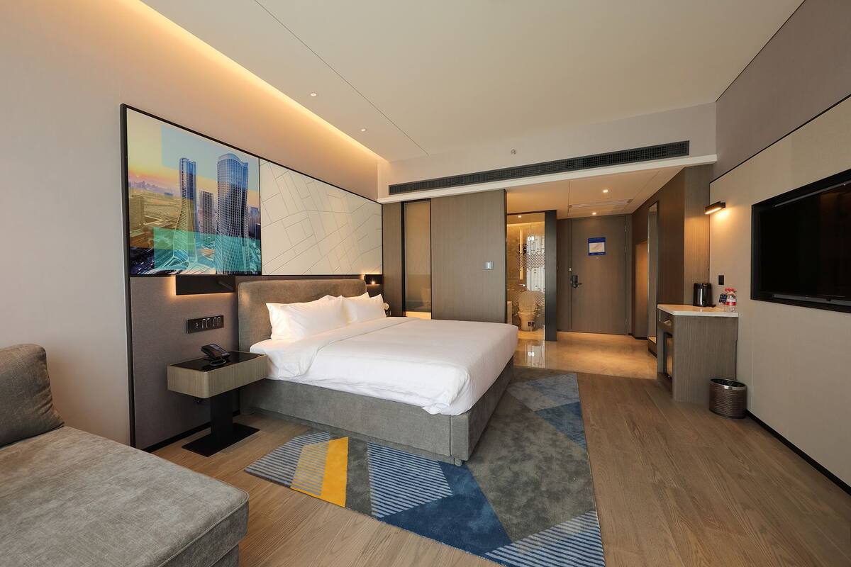 Habitación de hotel moderna con una cama grande, una mesita de noche con un teléfono, un televisor montado en la pared y un baño visible a través de una puerta abierta.