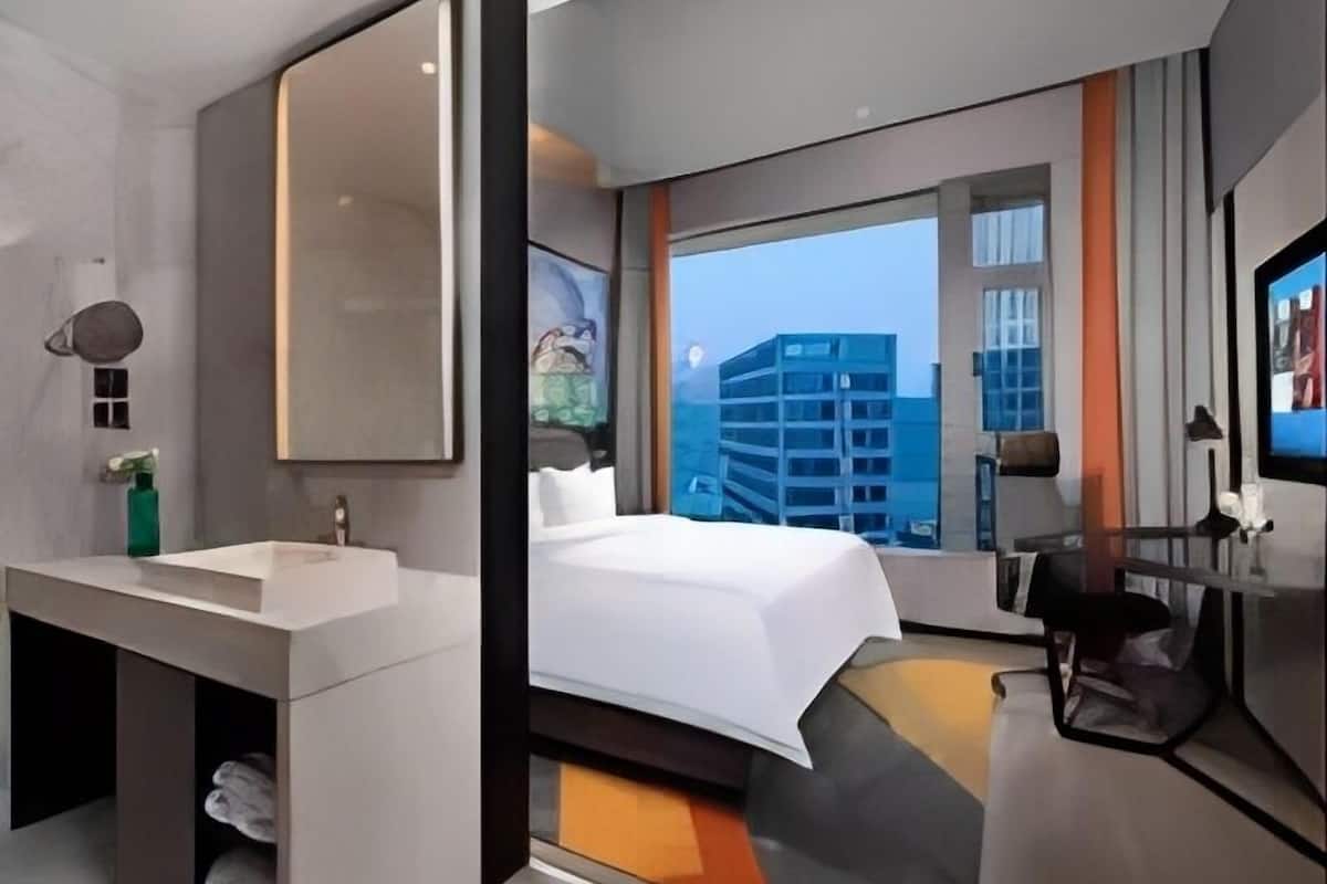 Una habitación de hotel moderna con una cama grande, un tocador con espejo, un escritorio y vistas a un paisaje urbano a través de un ventanal amplio.