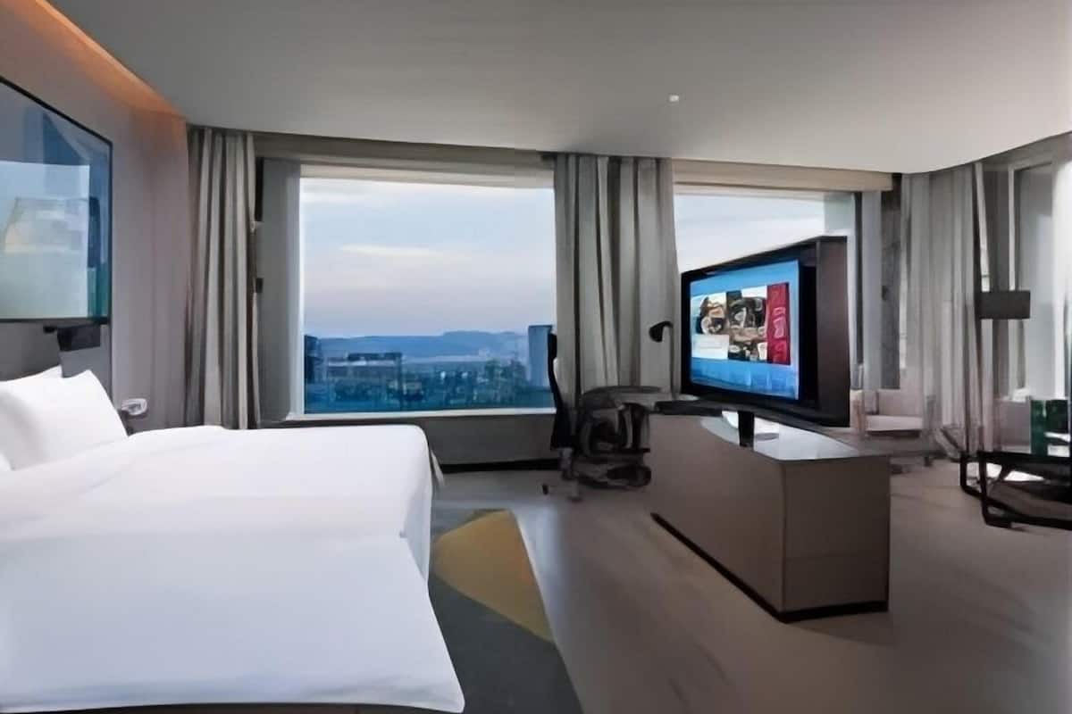 Una habitación de hotel moderna con una cama grande, un escritorio con televisión de pantalla plana, una silla y vistas de la ciudad a través de amplias ventanas.