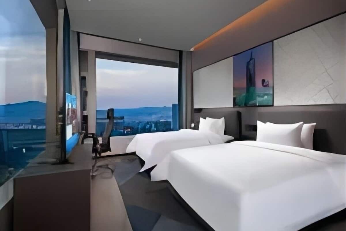 Una habitación de hotel moderna con dos camas, un ventanal grande con vistas a la ciudad y un televisor de pantalla plana.