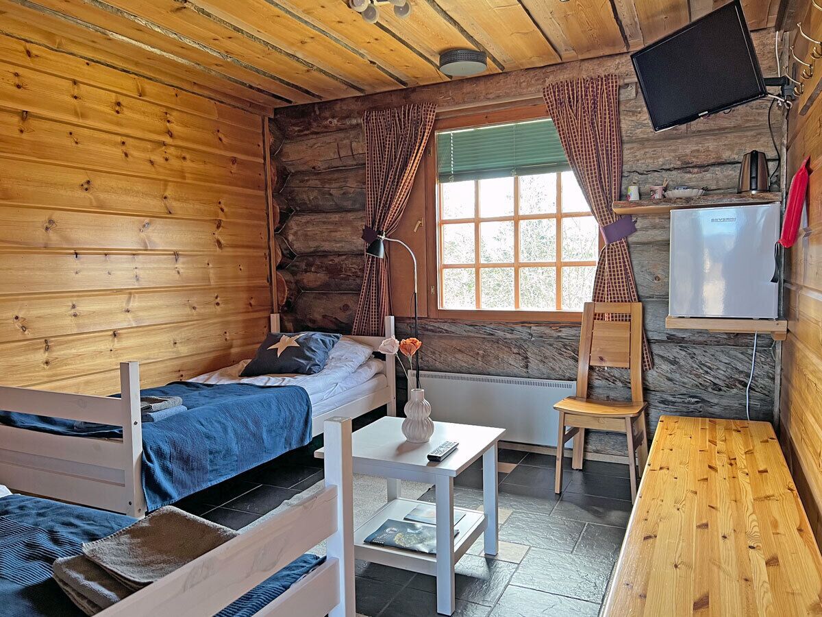 A cabin-style room with a bed, a small table with a vase, a chair, and a television mounted on the wall.