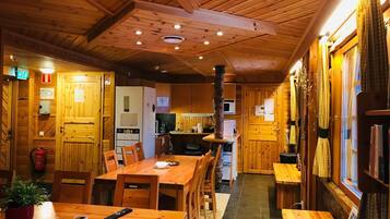 A cozy cabin interior with wooden walls and ceiling, a dining area with a table and chairs, a kitchen area with a refrigerator and microwave, and a sauna sign on the wall.