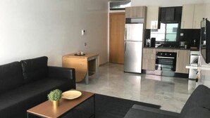 Un salon moderne avec un canapé noir, une table basse en bois et une petite table d’appoint. On aperçoit à l’arrière-plan une cuisine dotée d’appareils électroménagers en acier inoxydable et de meubles de rangement en bois clair.