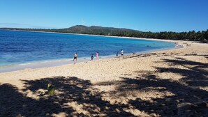 Una playa con agua azul cristalina, orilla de arena y aerogeneradores al fondo.