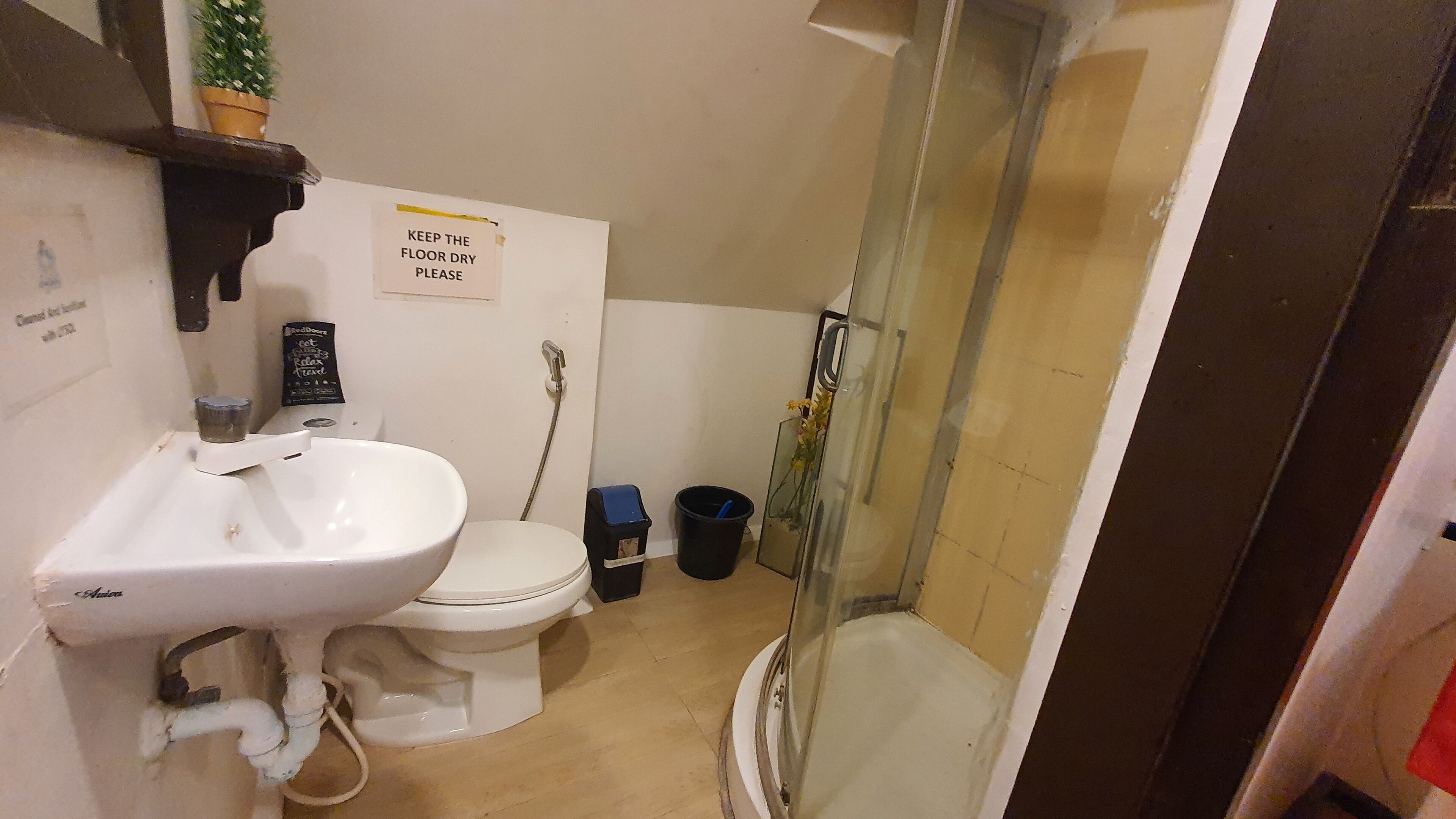 A bathroom with a white sink, toilet, and shower. There is a sign on the wall that says "KEEP THE FLOOR DRY PLEASE."