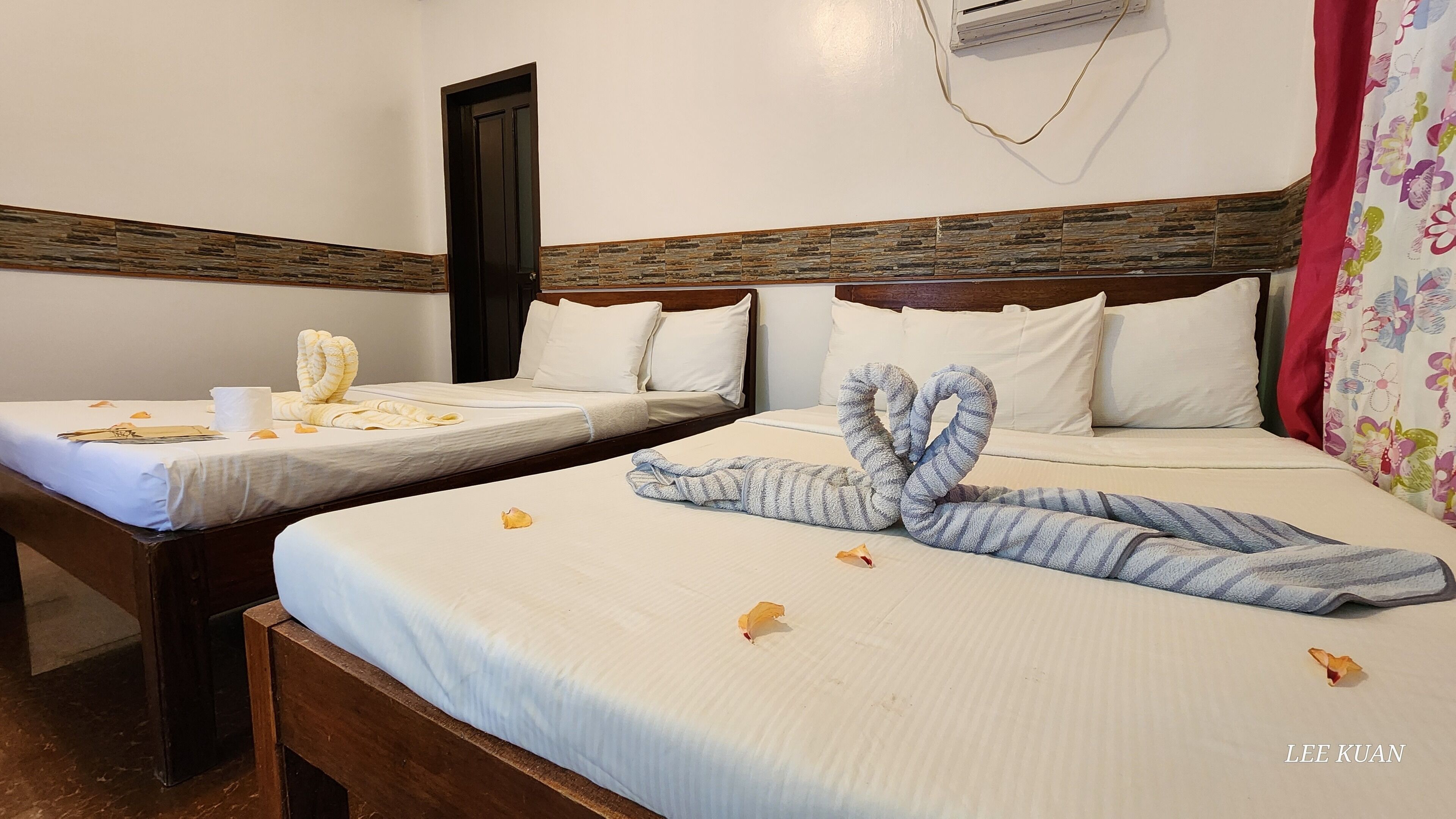 A hotel room with two beds, each with neatly made bedsheets and pillows. There is a wooden headboard, a wall-mounted air conditioning unit, and a floral curtain.