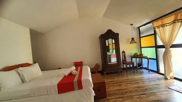 A bedroom with a bed, a wooden nightstand, a mirror, a table with chairs, and a view through a window with curtains.