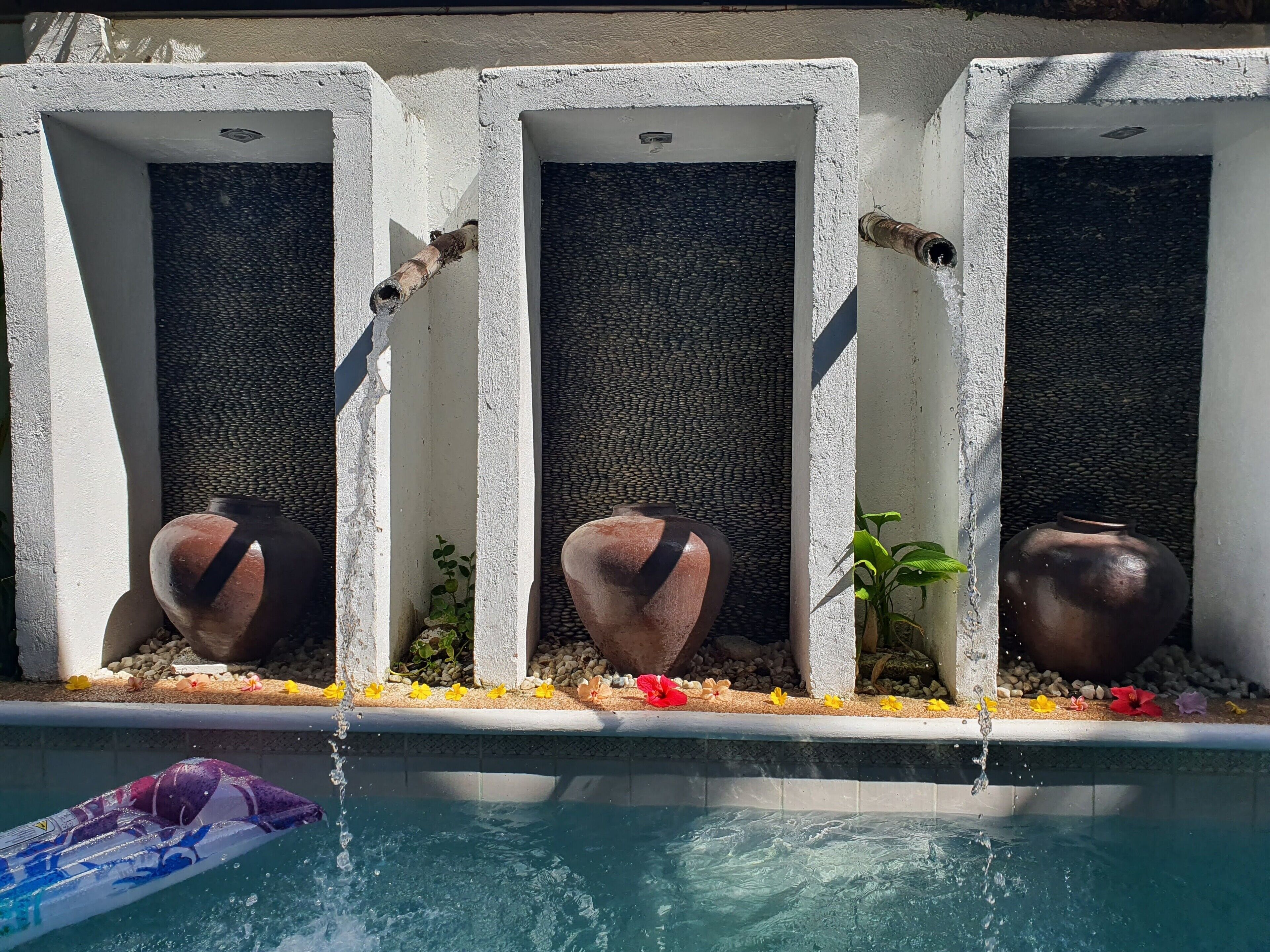 A pool with three concrete planters containing pots and small plants, surrounded by pebbles and flowers.
