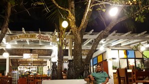 Un café frente a la playa con un letrero que dice "EVANGELINE Beach Resort", sillas de madera y un árbol grande en primer plano.