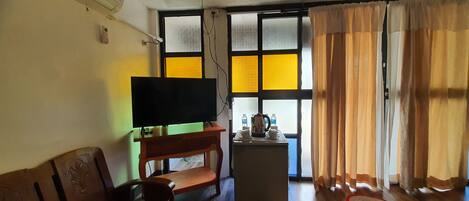 Ein Wohnzimmer mit Fernseher auf einem Holzboden, einem kleinen Tisch mit Wasserkocher und Tassen, einer Couch und einem Fenster mit Vorhängen.