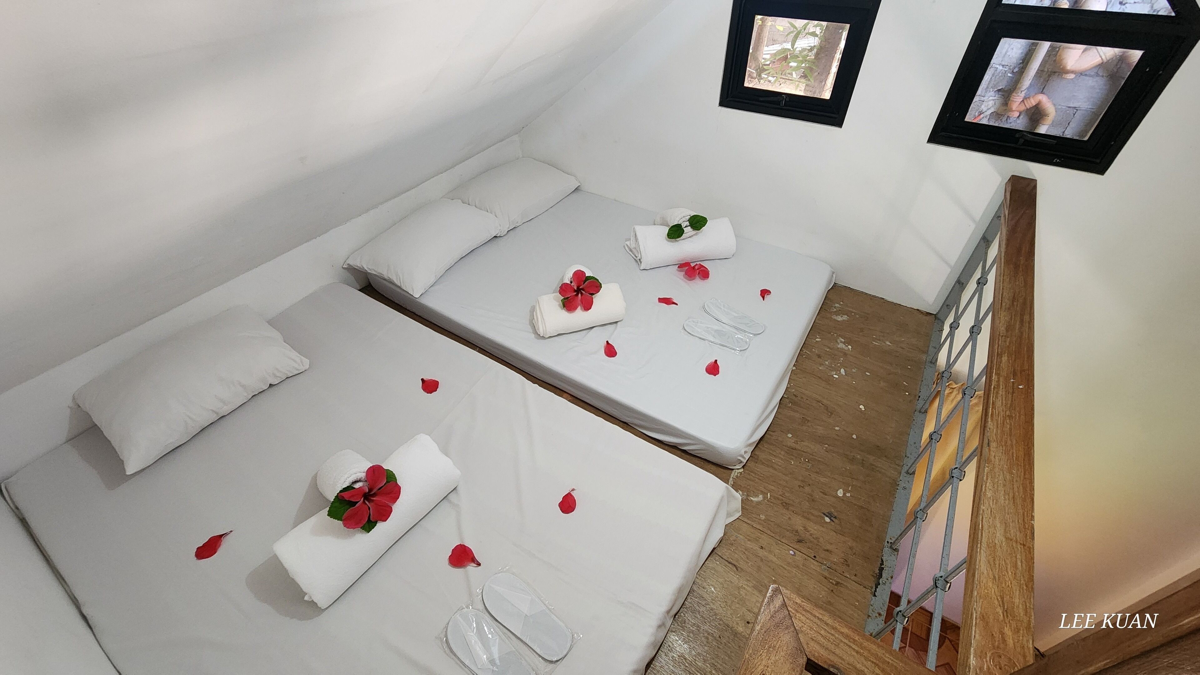 Une chambre avec deux lits, chacun recouvert de draps blancs et parsemé de pétales de fleurs rouges. Des tableaux ornent le mur, et une échelle en métal se trouve à côté des lits.