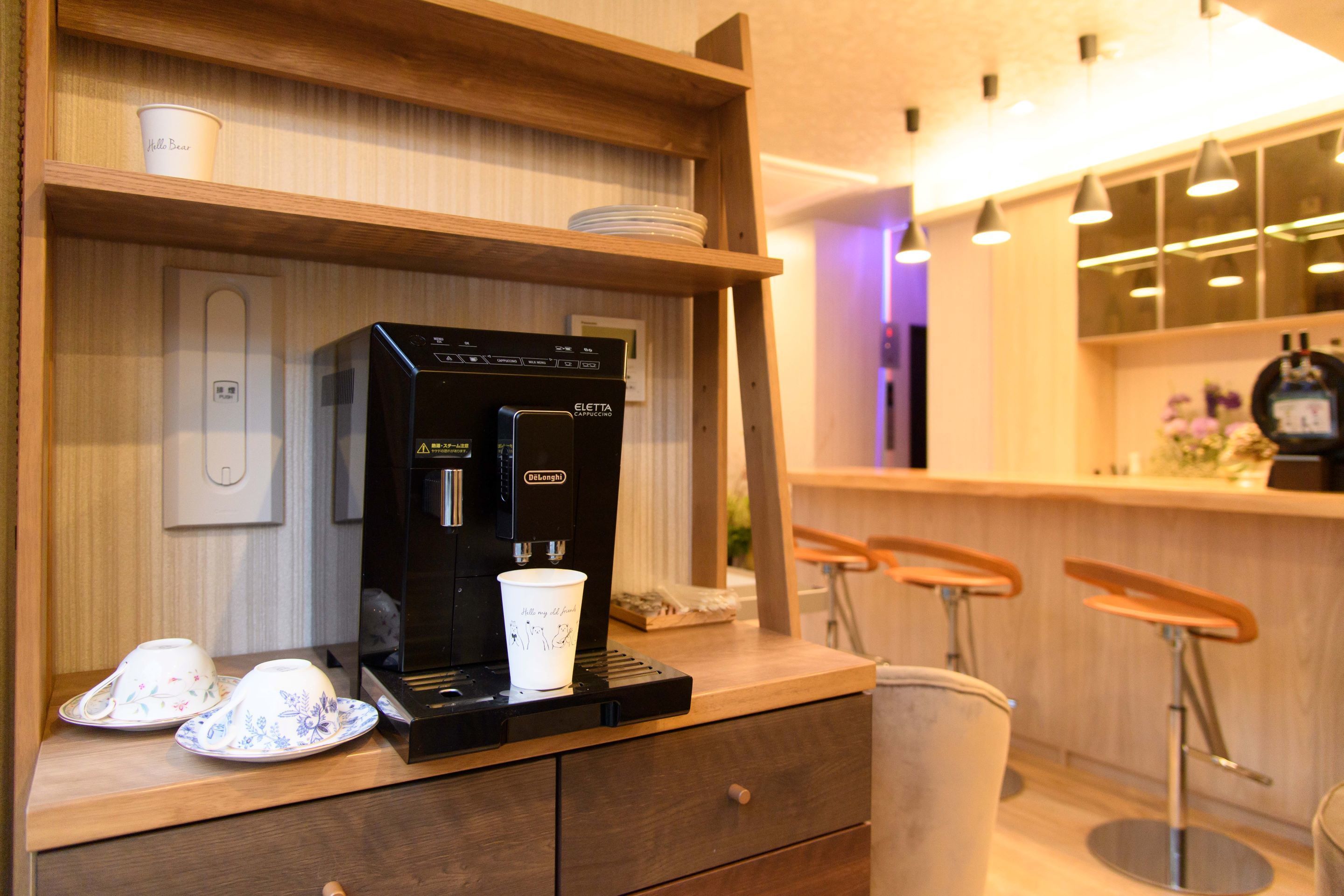 A coffee machine on a counter with a cup and saucer, a small trash bin, and a wooden shelf with a cup and plates.