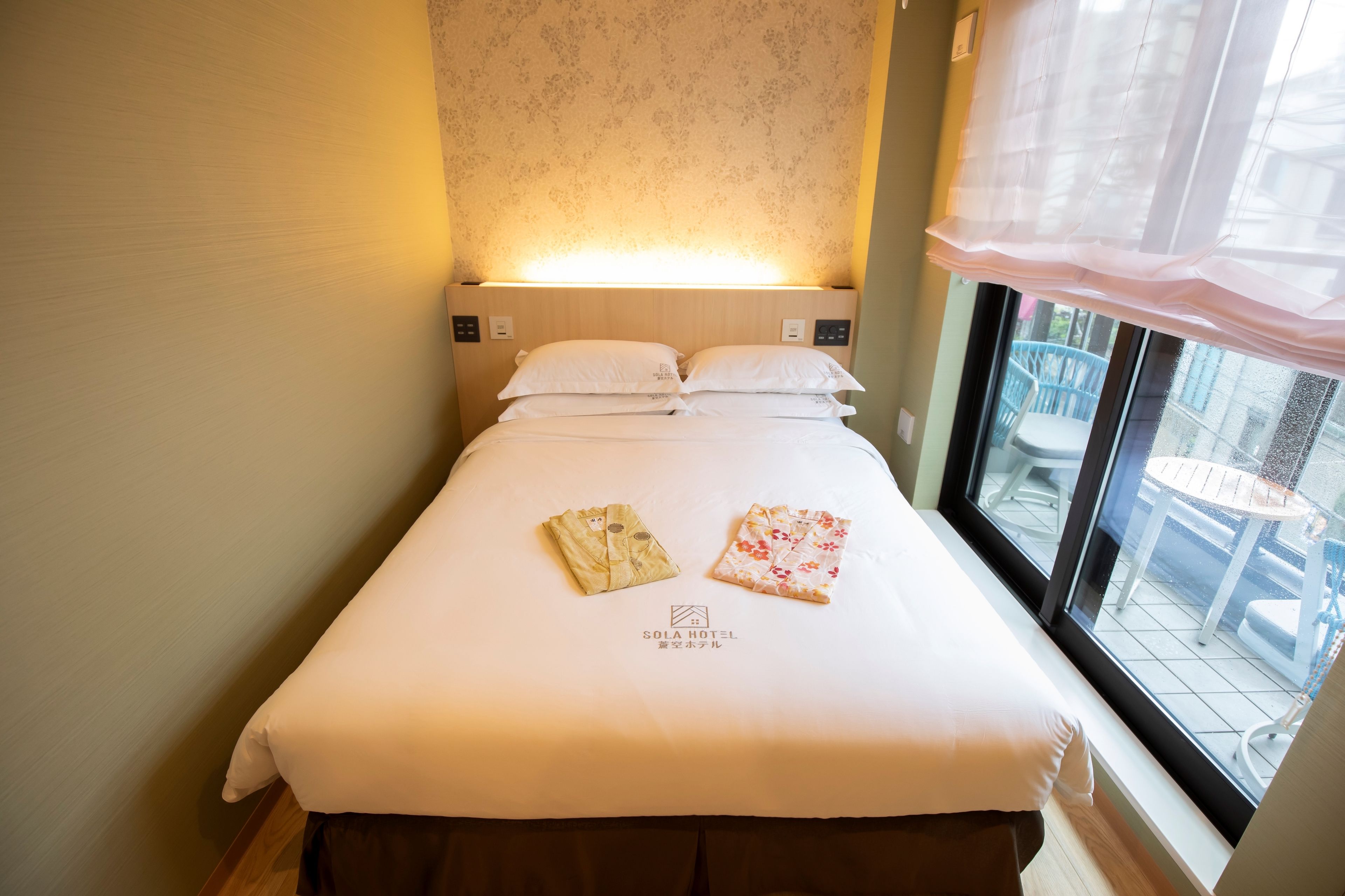 Habitación de hotel con una cama bien hecha, una ventana con vistas al exterior y un panel de control fijado a la pared.