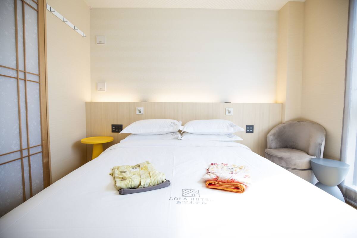 Une chambre d’hôtel avec un lit, des oreillers, une chaise, une petite table et une fenêtre.