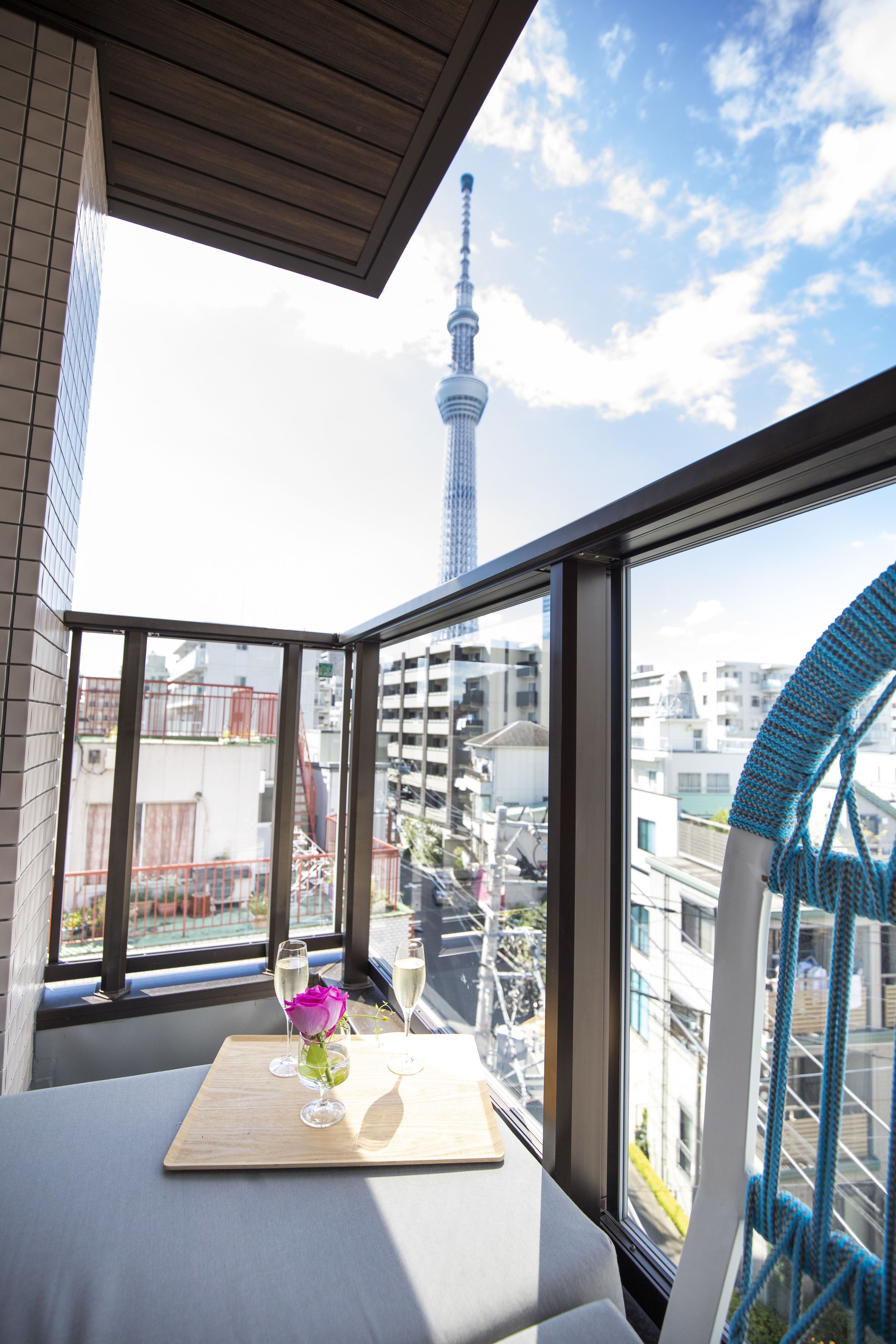 A balcony with a view of a tower, a table with a vase and glasses, and a blue rope structure.