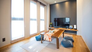 A modern living room with a large TV, wooden flooring, and large windows.