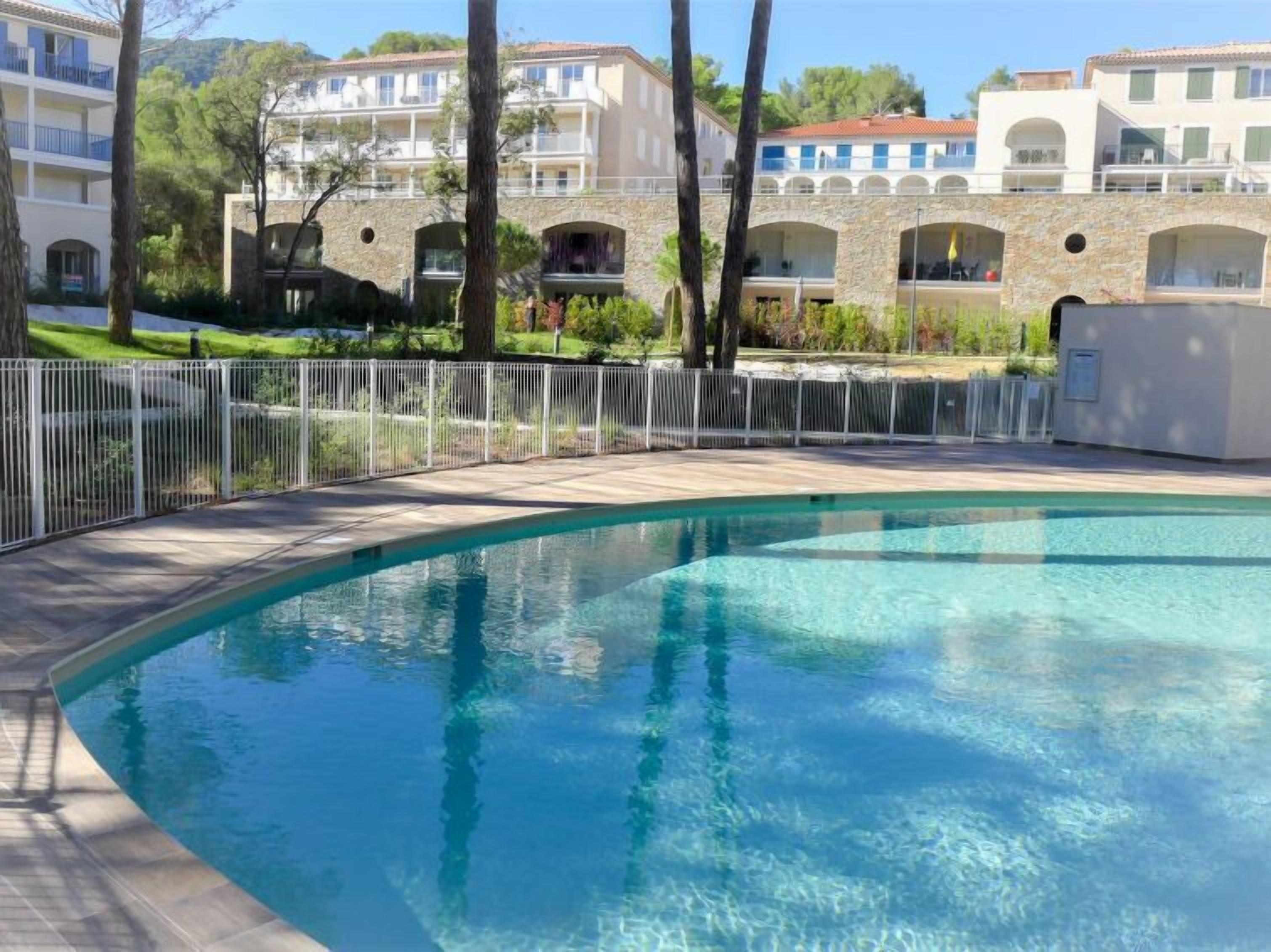 A swimming pool with a white fence, surrounded by buildings with balconies and a stone wall.