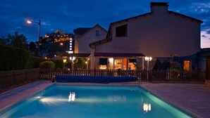 A swimming pool with a wooden deck, a covered patio area, and a building with a lighted entrance. - Les Messageries (Saint-Flour)