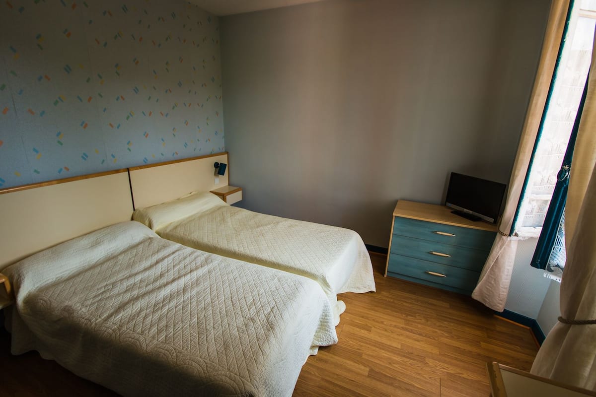 A room with two beds, a TV, a small dresser, and a window with curtains.