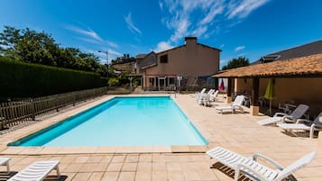 A rectangular swimming pool with a tiled deck, surrounded by white lounge chairs and a small covered seating area.