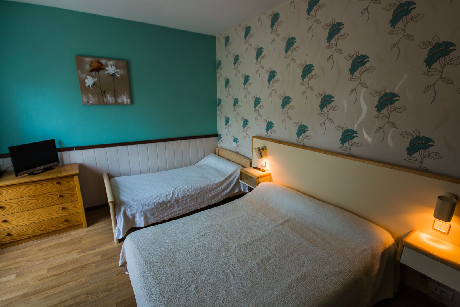 A room with two beds, a wooden dresser, a television, and a wall with floral wallpaper.