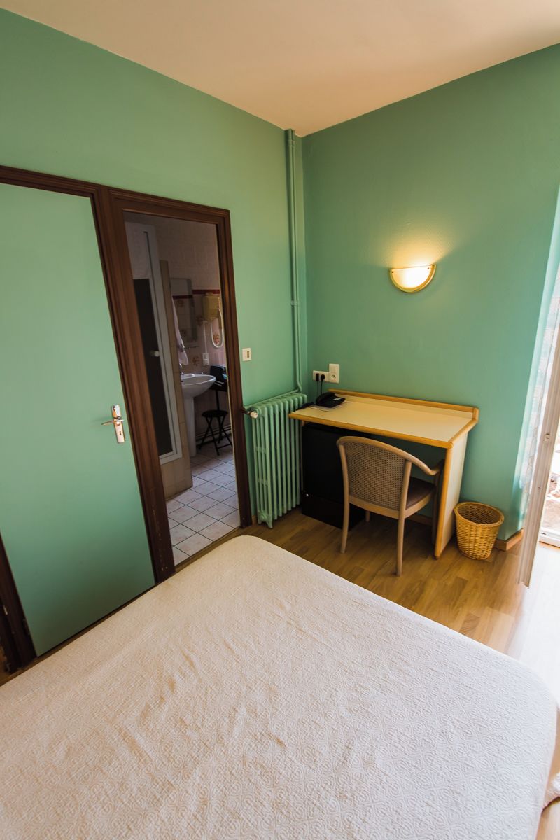 A small room with a bed, a desk, a chair, a radiator, and a light fixture.