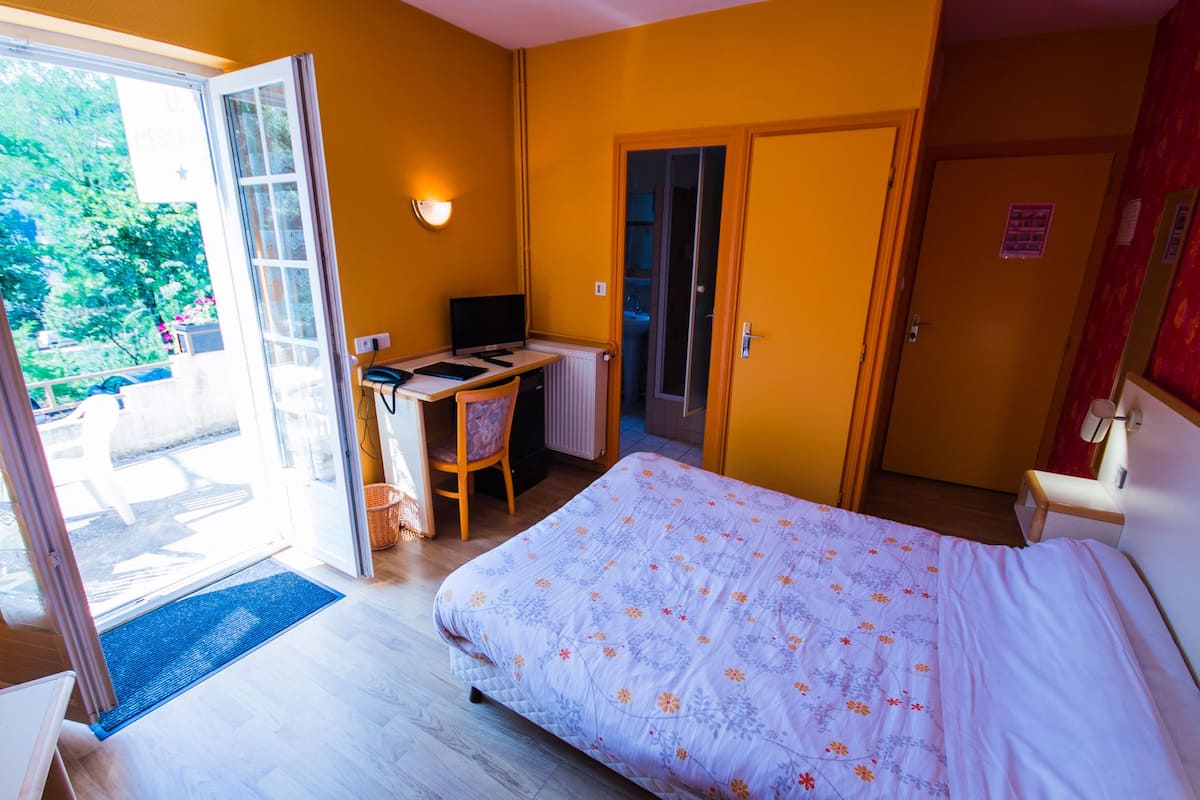 A bedroom with a bed, a desk, and a computer. There is a door leading to a bathroom and a view of the outside.