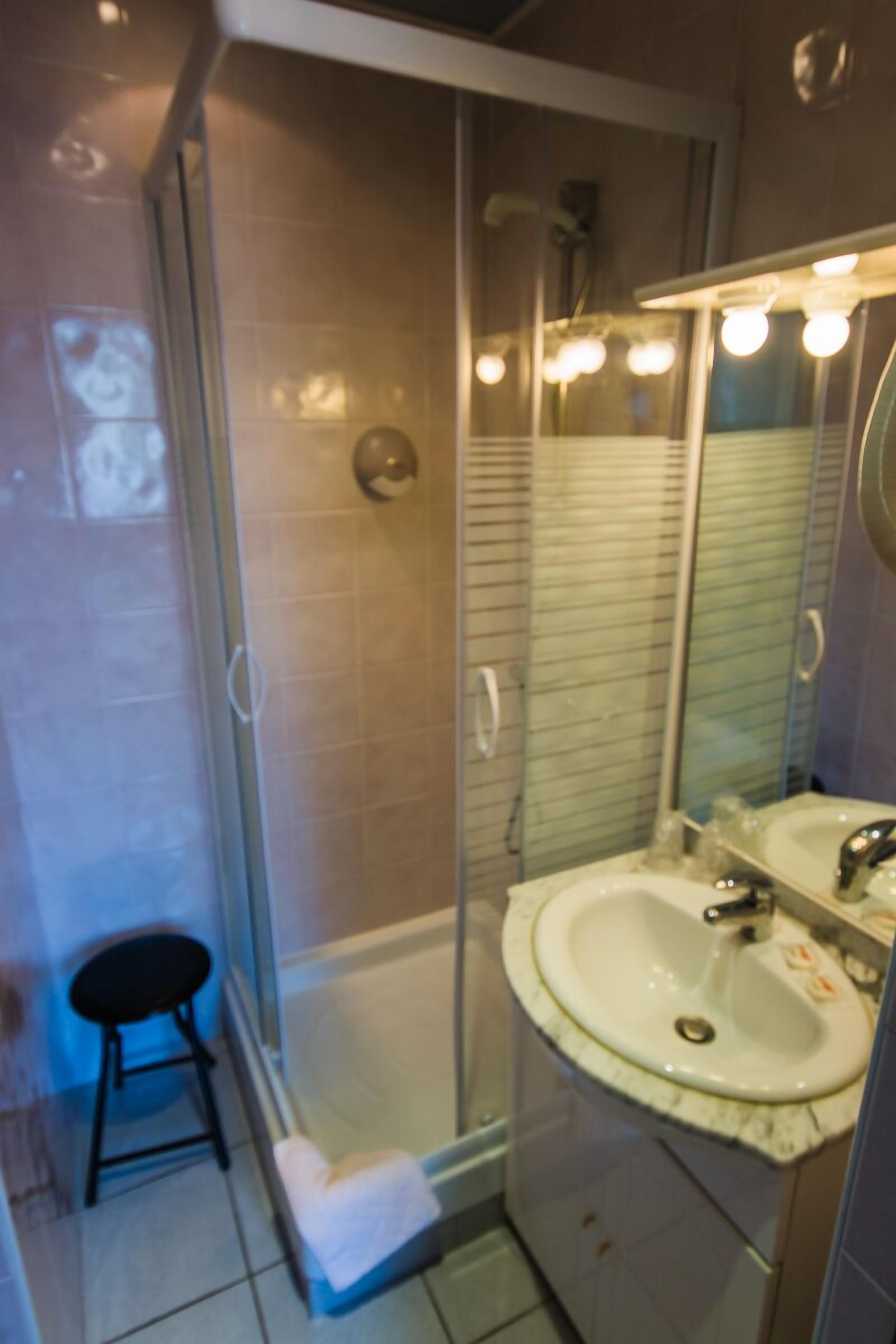 A bathroom with a glass-enclosed shower, a sink with a mirror, and a small stool.
