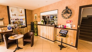 Un vestíbulo de hotel con mostrador de recepción, una zona de estar con dos sillones, una planta en maceta y un reloj de pared.