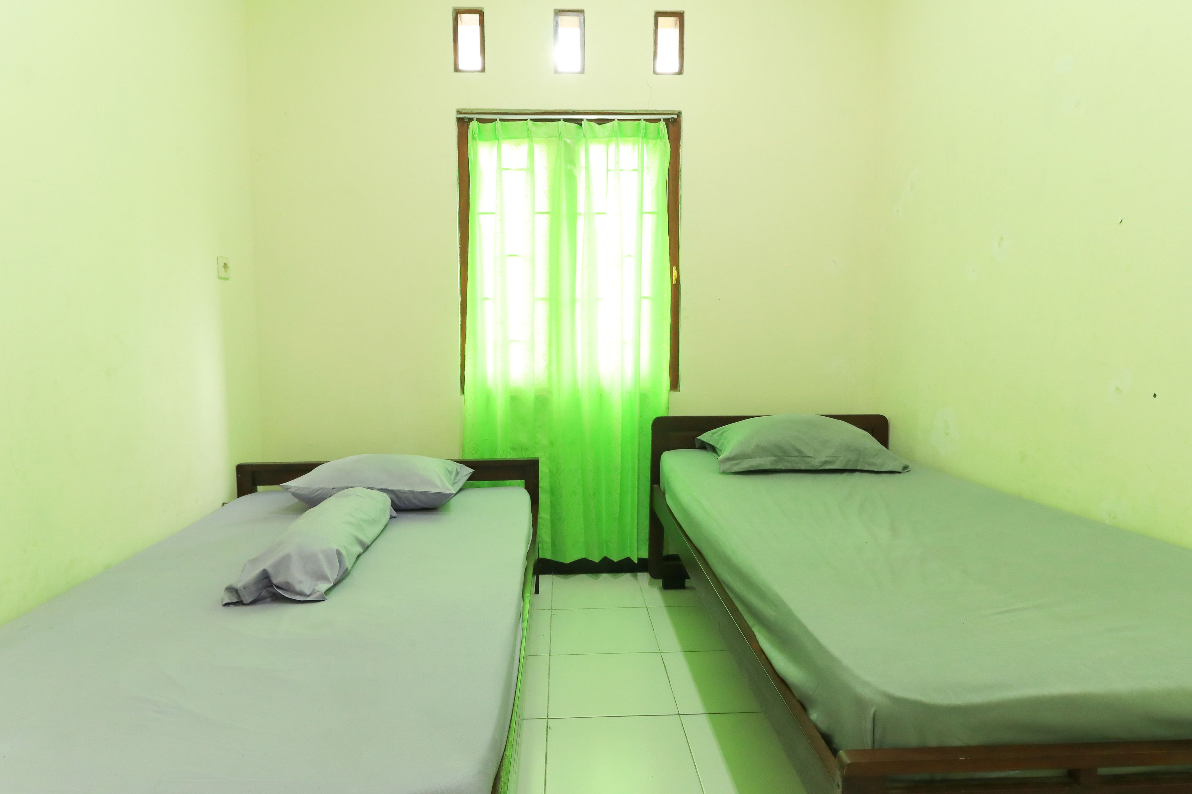 A small room with two single beds, a window with green curtains, and a light switch on the wall.