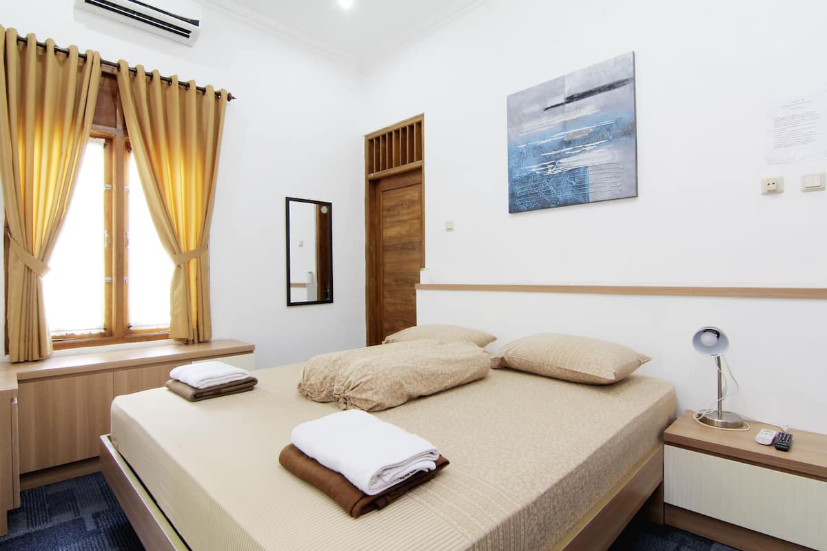 A hotel room with a bed, a nightstand, a window with curtains, a painting on the wall, and a wooden headboard.
