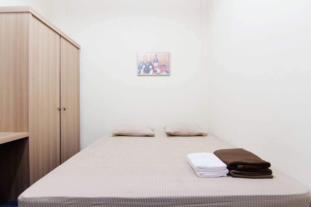 A single bed with a striped bedspread, two pillows, and a folded brown blanket on the right side. A wooden wardrobe is to the left of the bed.