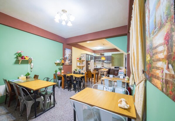 A dining area with multiple tables and chairs, a kitchen in the background, and a colorful painting on the wall. - Moonlight Fengqing Homestay (Xincheng)