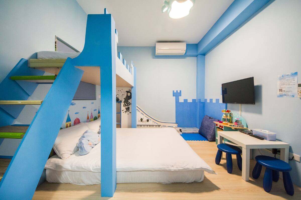 A child's bedroom with a bunk bed, a desk with stools, a TV, and a castle-themed wall decoration.