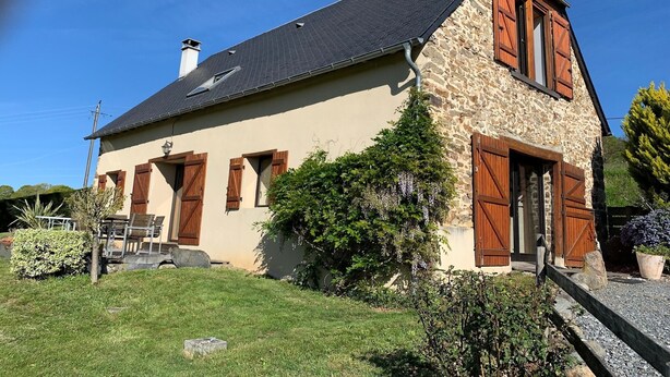 Ein Steinhaus mit geneigtem Dach und Holzläden, umgeben von einem gepflegten Rasen und verschiedenen Sträuchern.